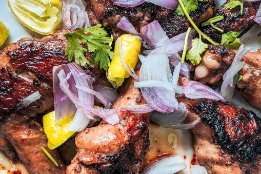 Grilled meat pieces topped with lemon wedges, sliced red onions, herbs, and spices in a savory sauce, ready to eat.