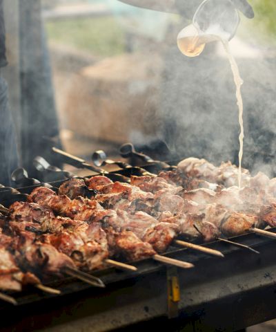 Smoky outdoor grill with skewered chicken and meat as oil pours overhead and flames rise over the coals.