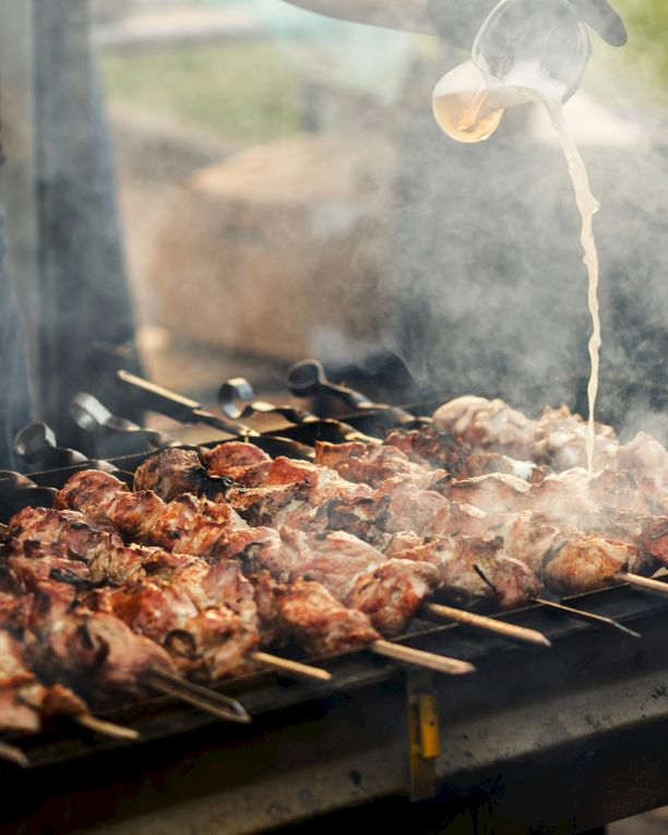 Smoky outdoor grill with skewered chicken and meat as oil pours overhead and flames rise over the coals.